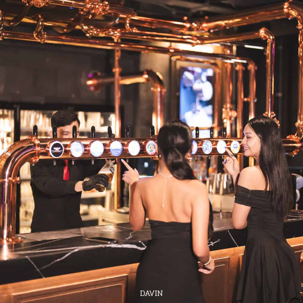 A bartender serving a glass of draft beer to two women at a bar with an elaborate copper tap system.