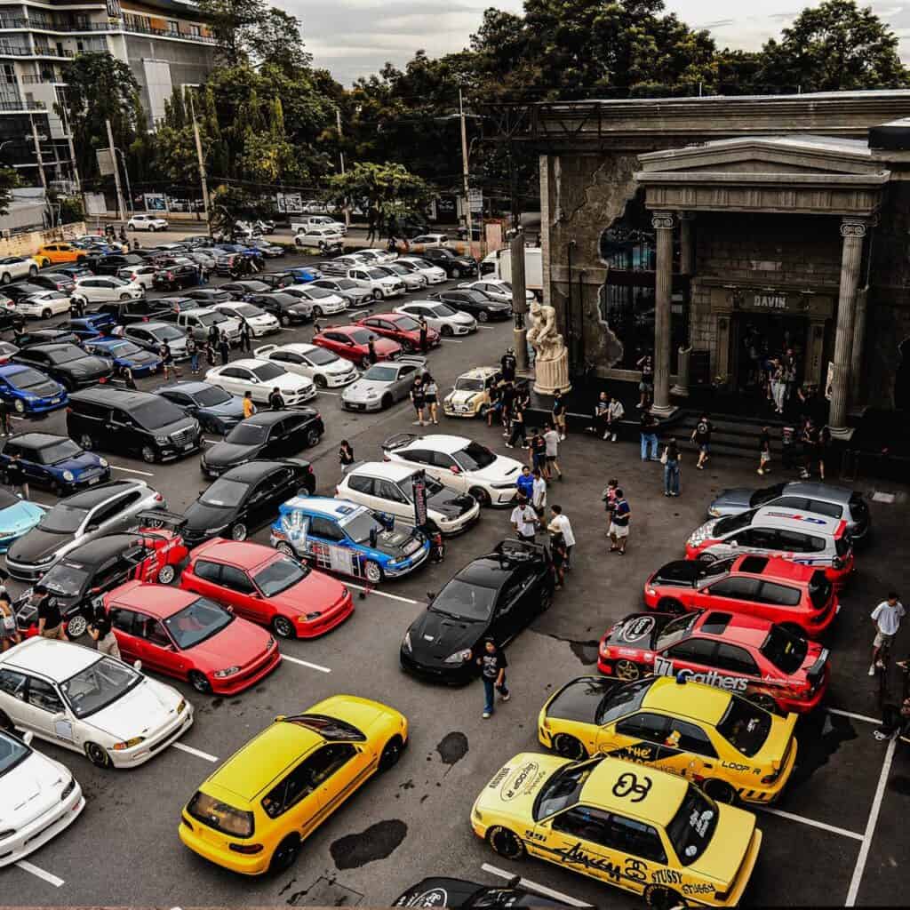 An aerial view of a large car meet with many modified cars and people gathered in front of the Davin Cafe building.