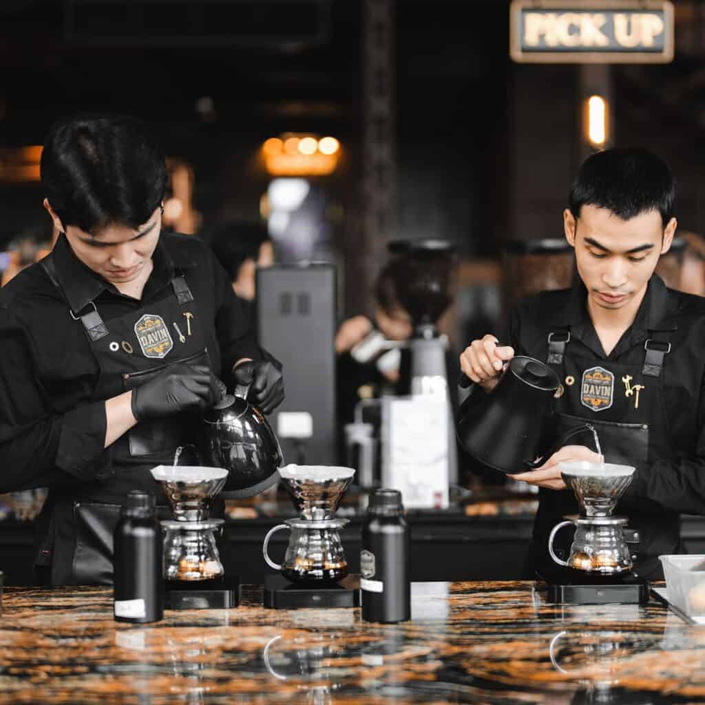Two Davin Cafe baristas meticulously preparing pour-over coffee using gooseneck kettles at a counter.
