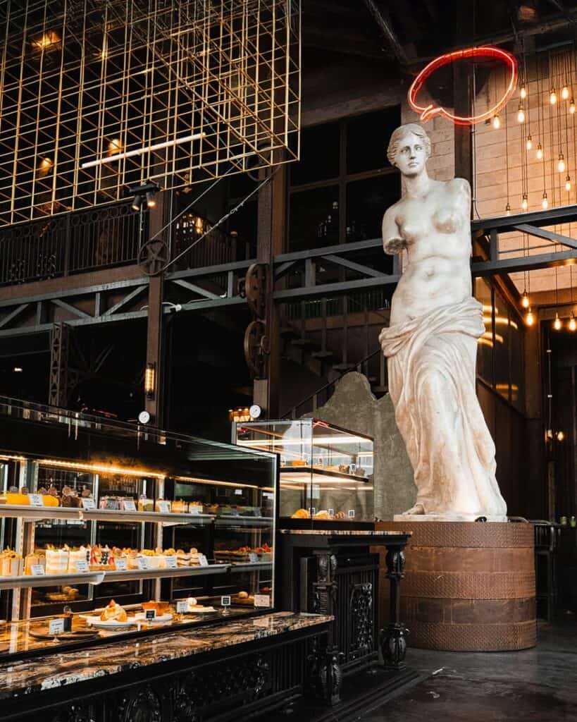 รูปปั้นวีนัส (Venus di Milo) ขนาดใหญ่ตั้งอยู่ข้างตู้โชว์เค้กที่ส่องสว่าง และโครงสร้างเหล็กสีทองภายใน Davin Cafe คาเฟ่เปิดดึก.