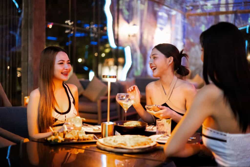 Three women laughing and celebrating while enjoying food and drinks at a brightly lit table inside the modern banquet restaurant at night.
