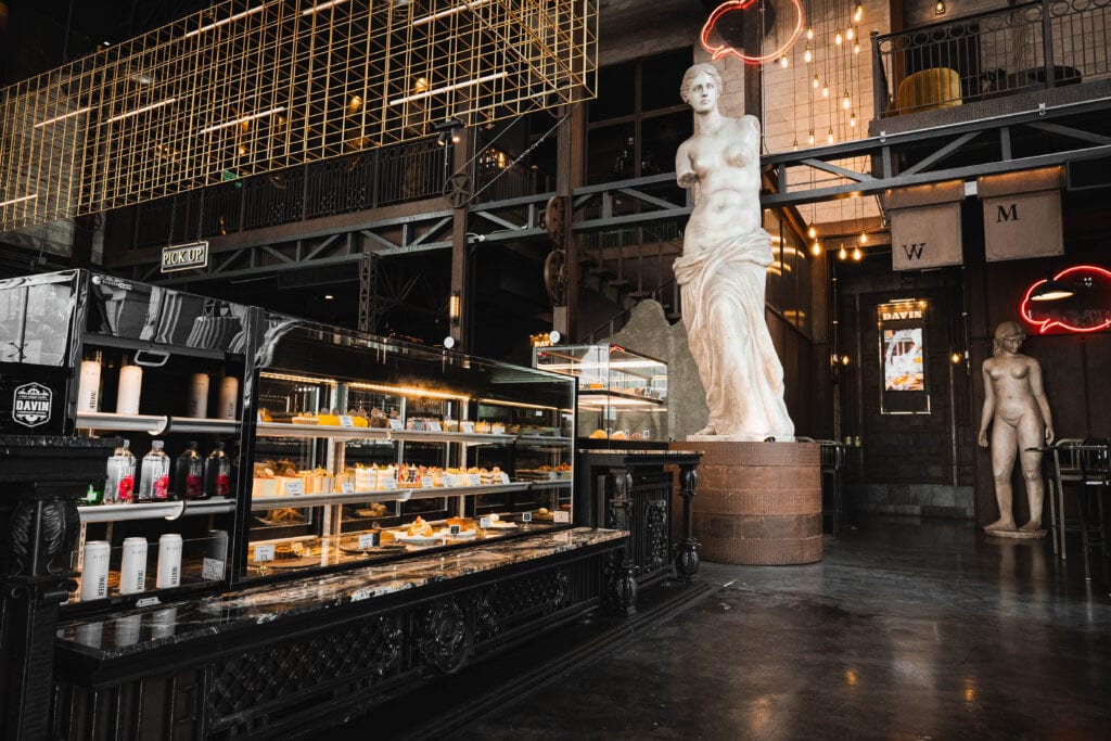 โซนเคาน์เตอร์ขนมและเบเกอรี่ภายในคาเฟ่รามอินทรา ตกแต่งสไตล์อินดัสเทรียลหรู ผสานประติมากรรมคลาสสิกและโครงเหล็กสีทอง สร้างบรรยากาศโดดเด่นให้ Davin Cafe เป็นจุดนัดพบและถ่ายภาพยอดนิยม