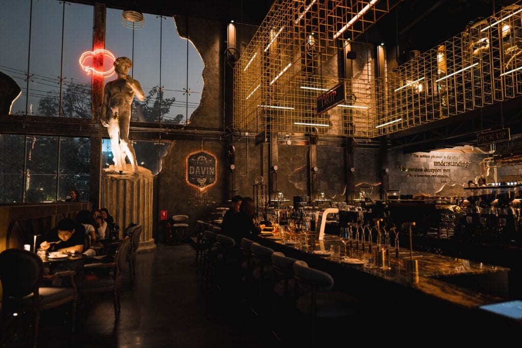 ร้านแฮงเอาต์บรรยากาศกลางคืน สไตล์อินดัสเทรียล มีเคาน์เตอร์บาร์ยาว ลูกค้านั่งดื่มและพูดคุยชิล ๆ เหมาะสำหรับแฮงเอาต์ยามค่ำคืน