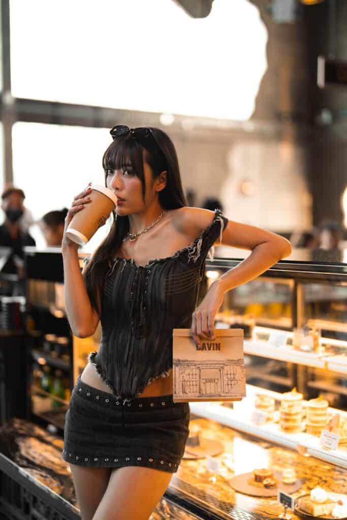 A stylish woman enjoying coffee inside a photogenic cafe with warm lighting and a dessert display, perfect for lifestyle photography, cafe hopping, and visual content creation
