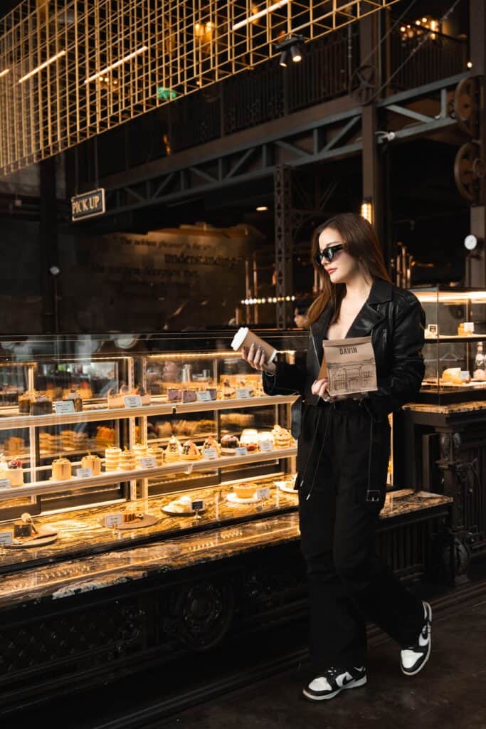 ลูกค้าผู้หญิงกำลังเลือกขนมและถือกาแฟหน้าตู้เบเกอรี่ของร้านกาแฟเลียบด่วน รามอินทรา บรรยากาศภายใน Davin Cafe สไตล์อินดัสเทรียลหรู ให้ฟีลคาเฟ่ไลฟ์สไตล์ที่ทั้งถ่ายรูปสวยและนั่งใช้เวลาได้จริง