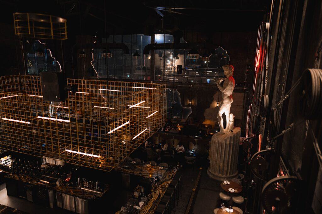 An elevated interior view of Davin Cafe, a Ramintra Expressway cafe featuring a dramatic industrial-style space with a classical statue centerpiece, warm ambient lighting, and a spacious nighttime atmosphere designed for both dining and visual experience.