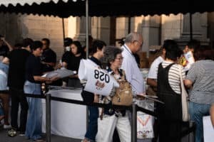 ผู้คนเข้าคิวลงทะเบียนงานอีเวนต์ที่ Davin Cafe ในบรรยากาศกลางแจ้ง, มีผู้หญิงและผู้ชายหลายคนรอรับบัตรเ.