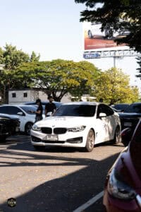 ที่จอดรถยนต์ในสวนสาธารณะกลางแจ้งในช่วงบ่าย, รถยนต์สีขาวจอดอยู่ในพื้นที่จอดรถ, ต้นไม้และป้ายโฆษณาในพื.