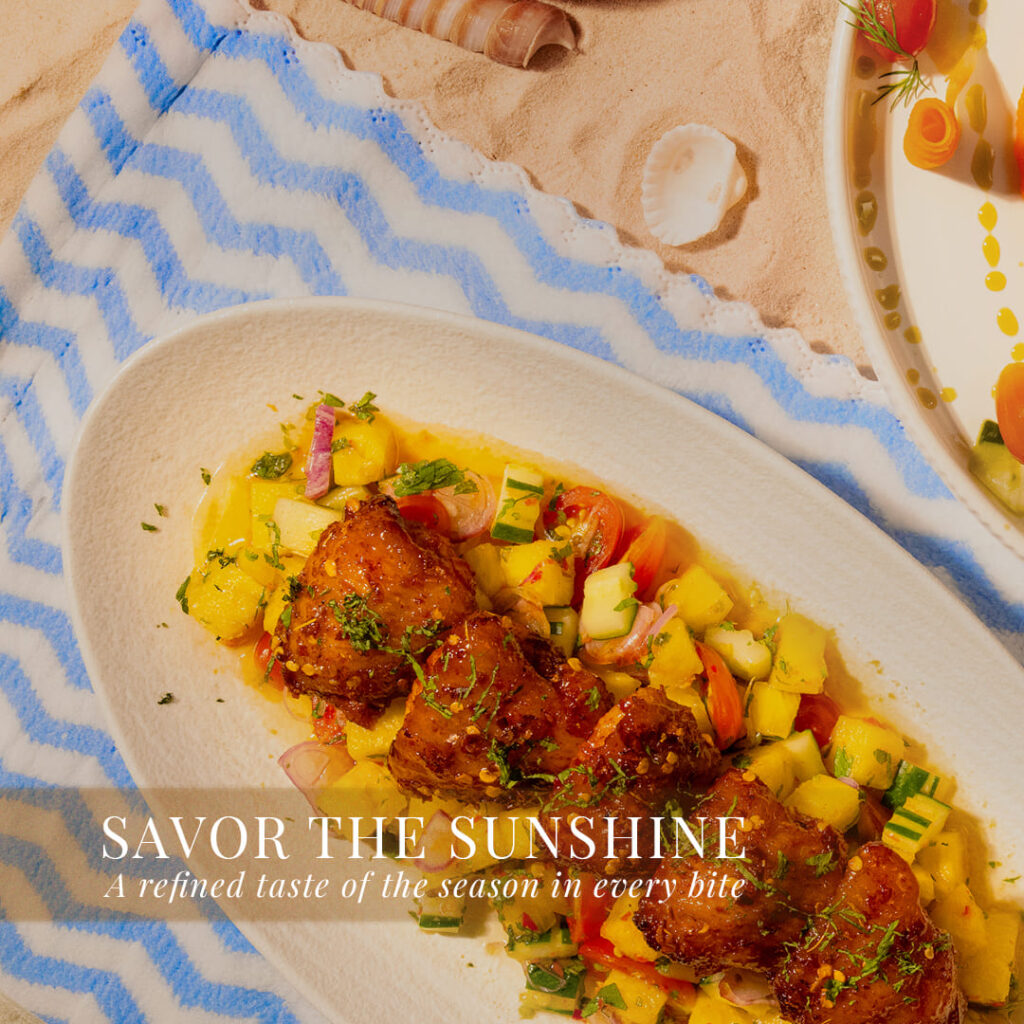 Davin's chicken spicy sweet and sour, a vibrant fusion summer menu main course, presented on beach sand next to fresh pineapple and miniature umbrellas, offering a refreshing and unique taste experience this season.