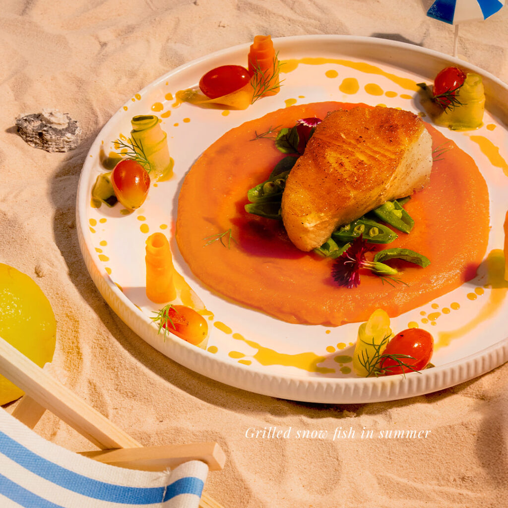 Davin's grilled snow fish with a vibrant orange summer sauce and garnish, a premium seafood summer menu main course, creatively presented on sand to elevate the high-end summer dining experience on Pradit Manutham Road.