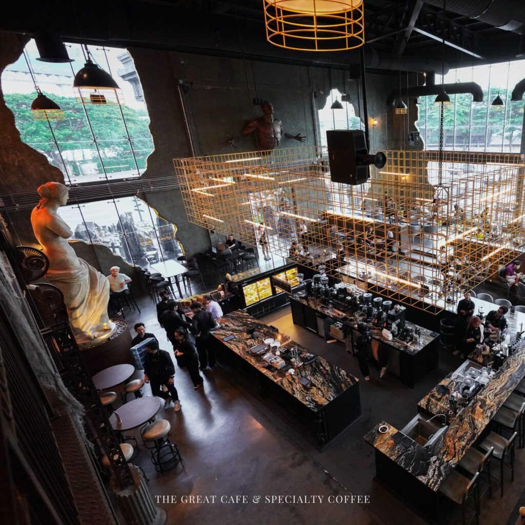 A high-angle view of the grand interior at Davin Cafe, a premium coffee shop featuring a massive specialty coffee bar, industrial loft design, and a striking classical Greek sculpture.