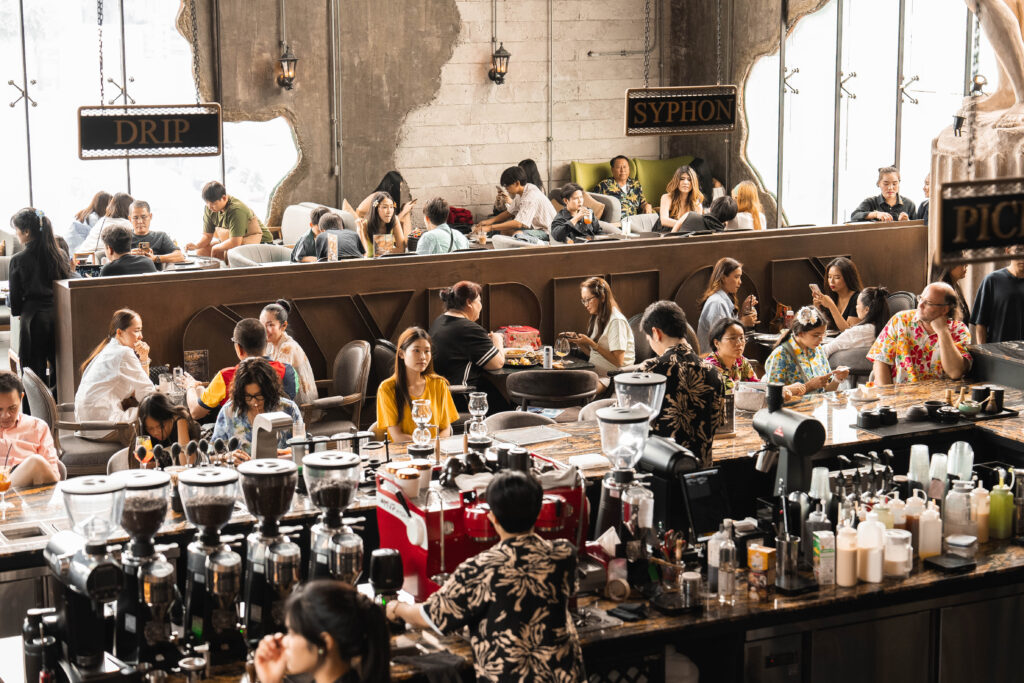 A vibrant indoor scene at Davin Cafe during the Songkran festival, with guests filling the coffee bar and seating areas, offering a comfortable urban escape from the heat in Bangkok