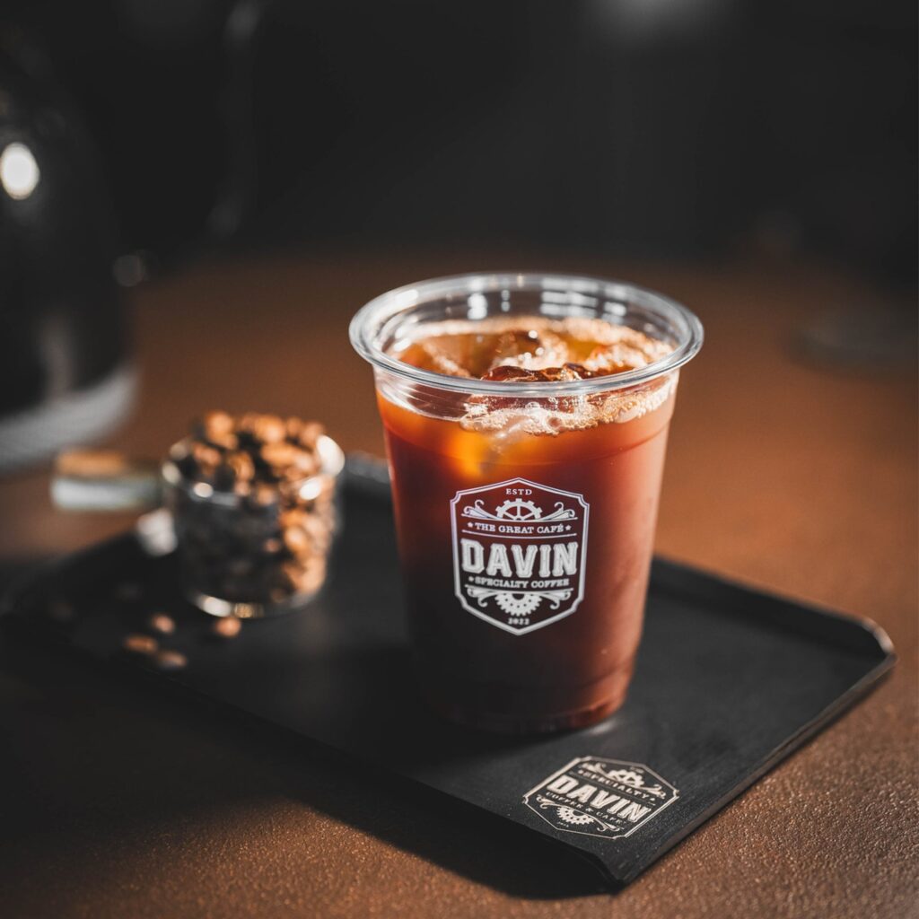 กาแฟเย็นในแก้วใสของ Davin Cafe วางบนถาดพร้อมเมล็ดกาแฟด้านหลัง สื่อถึงประสบการณ์การดื่ม กาแฟ คั่วอ่อน ที่ให้รสชาติใส สดชื่น และดึงกลิ่นโน้ตผลไม้ได้ชัดเจน