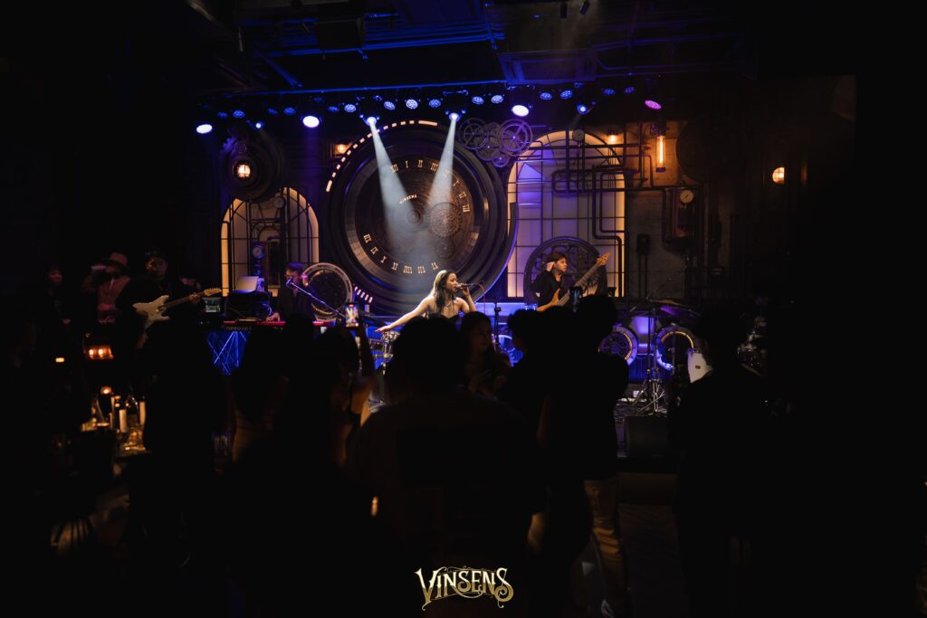 บรรยากาศดนตรีสดสุดมันภายในร้าน Vinsens ร้านนั่งชิล รามอินทรา ผู้คนกำลังสนุกกับเสียงเพลงและแสงไฟบนเวที สะท้อนประสบการณ์แฮงเอาท์ยามค่ำคืนที่เต็มไปด้วยพลัง