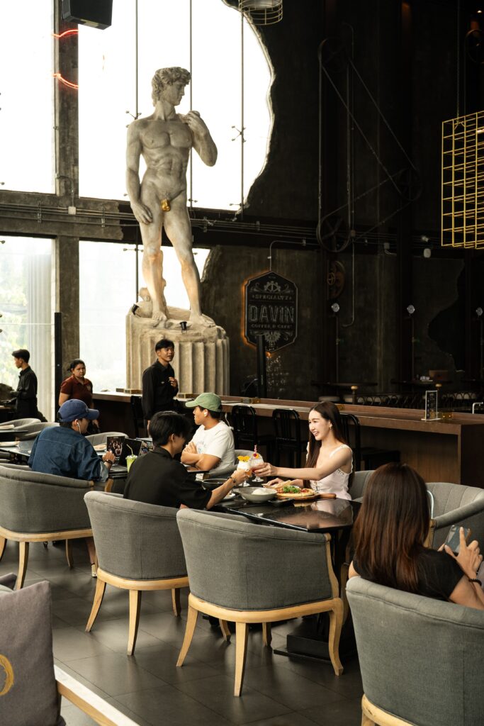 ลูกค้ากำลังรับประทานอาหารและเครื่องดื่มภายใน DAVIN CAFE ร้านอาหาร เลียบด่วน บรรยากาศหรูเพดานสูง พร้อมแลนด์มาร์กประติมากรรมคลาสสิก เหมาะสำหรับมื้อพิเศษ การสังสรรค์ และประสบการณ์อาหารระดับพรีเมียม